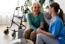 Oudere vrouw in groen shirt zit op de bank, met achter haar haar rollator. Zorgmedewerker zit naast haar op de bank, houdt haar ene hand vast en heeft haar andere hand op de schouder van de vrouw. Ze lachen naar elkaar en zijn aan het praten.